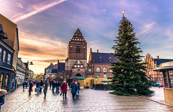 A busy street at dusk featuring people, a large Christmas tree, a historic building, and a sky with pink and blue hues.