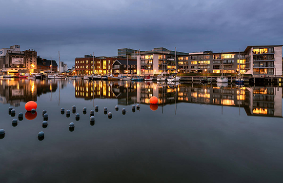 A calm evening view of a waterfront with modern buildings and docked boats, reflecting on the water surface, under a dusky sky.