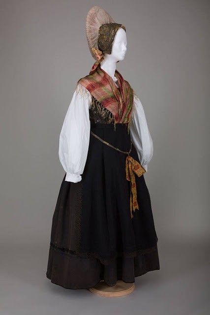 A mannequin is displayed in traditional folk attire, including a white blouse, black skirt, vest, and patterned scarf.