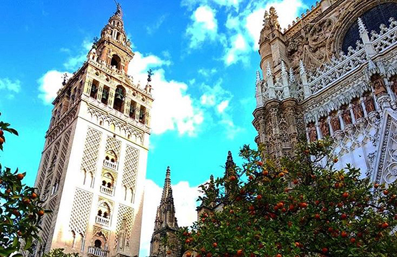 A photo captures the detailed architecture of Seville Cathedral's Giralda bell tower, set against a blue sky with greenery and orange trees.