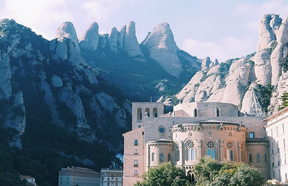 A historic, ornate building with jagged mountains behind it under a lightly clouded blue sky.