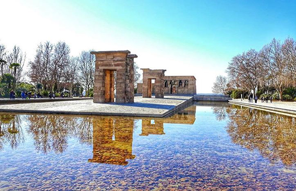 A picturesque view of the Temple of Debod, its Egyptian architecture mirrored in water, under a blue sky with flanking trees.