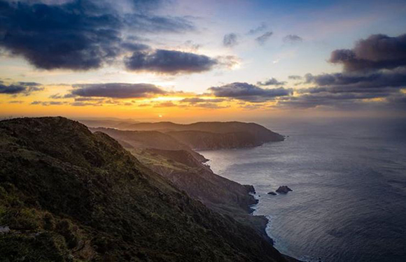 A scenic coastal landscape at sunset with rolling hills and the ocean extending towards the horizon under a sky with clouds.