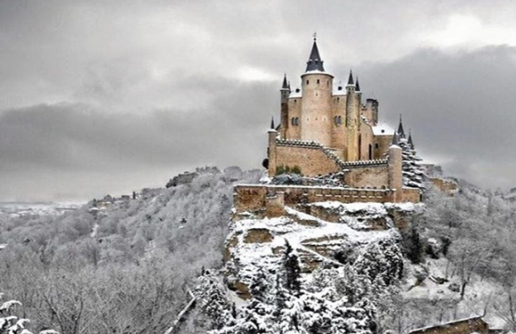 A castle with multiple towers and turrets perched on a hill, surrounded by trees and landscape covered in snow. The sky is overcast.