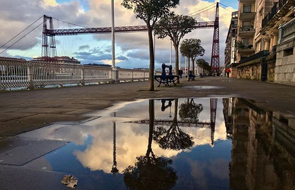 A cityscape with a large bridge in the background, a wet pavement reflecting the scene, and a few people walking along the sidewalk.