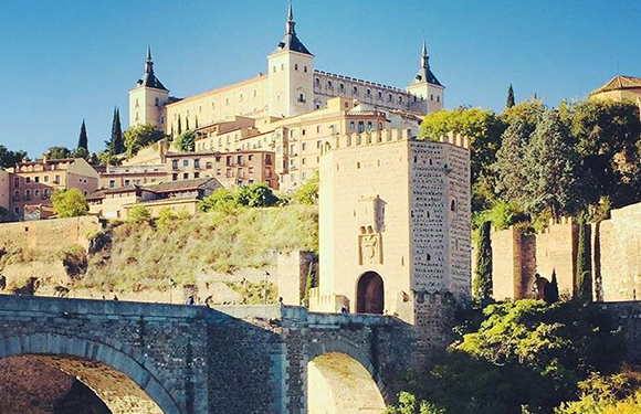 A historic castle on a hilltop with a stone bridge in the foreground, surrounded by greenery under a clear sky.