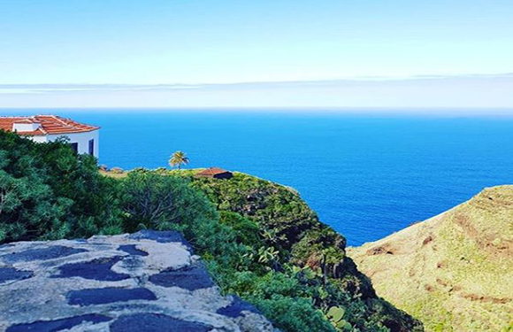 A coastal scene featuring a blue sky, distant ocean, a green cliff, and a building with a terracotta roof.