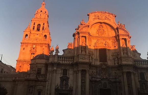 A photograph of a historic cathedral bathed in the warm glow of sunset, showcasing detailed architecture and a bell tower against a clear sky.