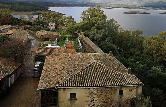 Aerial view of a rustic village with traditional stone buildings and tiled roofs near a body of water surrounded by greenery.