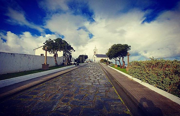 A cobblestone street leading towards a white church with a bell tower under a partly cloudy sky, flanked by greenery and a white fence on the left.