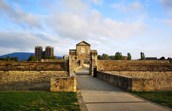 A stone bridge leading to an arched gate of a historical fortress with towers in the background under a partly cloudy sky.