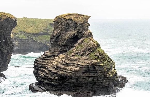 A coastal scene featuring a weathered rock, turbulent sea, distant cliffs, and an overcast sky.