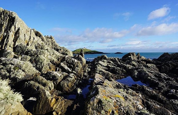 A coastal landscape with jagged rocks in the foreground and a small island in the distance under a blue sky with scattered clouds.