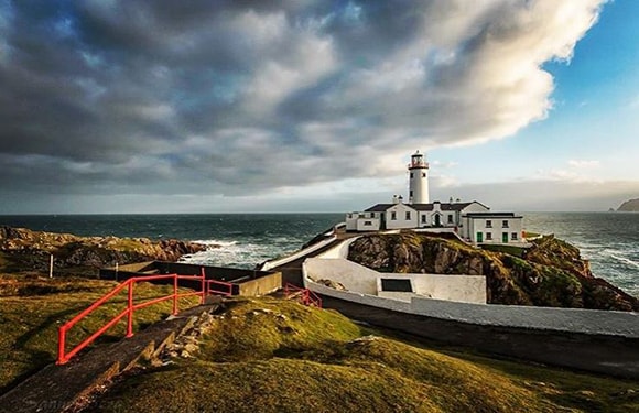 A picturesque lighthouse situated on a rugged coastline with a red handrail leading towards it, under a dramatic sky with clouds.