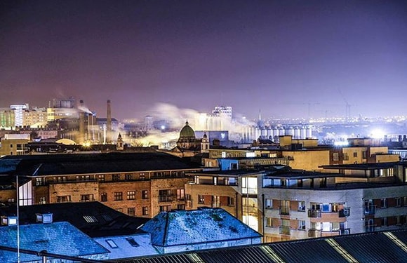 A night cityscape featuring industrial buildings and a brightly lit, steam-emitting factory under a dark sky.