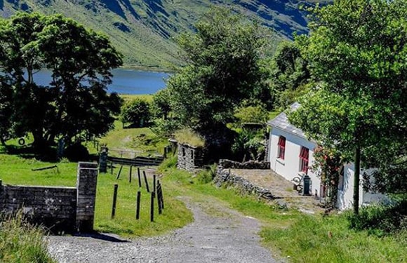 A small white house with a red door located beside a country road with a stone wall, surrounded by greenery with a lake and hills in the background.