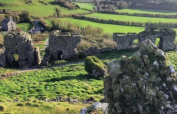 An old stone structure with archways sits in ruins amidst green grass, trees, fields, under a clear sky.