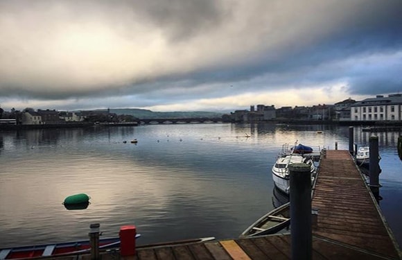 A peaceful waterfront view with a cloudy sky, calm water, a wooden dock, moored boats, and distant buildings.