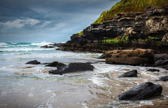 A rocky shoreline with waves crashing against the rocks under a cloudy sky.