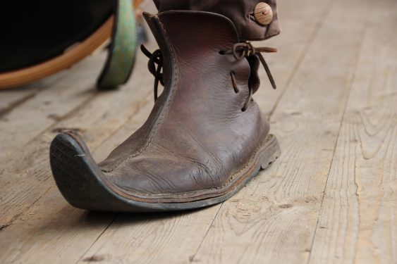 A single, worn brown leather boot with curled-up toe on a wooden floor.