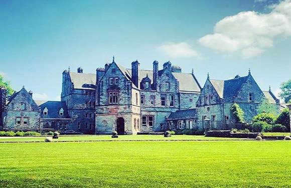 A large, historic stone mansion with multiple chimneys and turrets, surrounded by a well-manicured lawn and set against a clear blue sky.