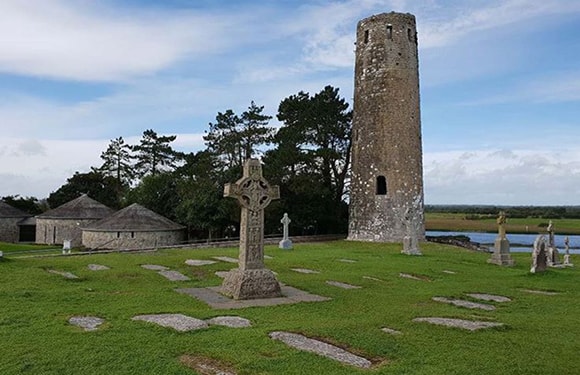 A historical site with an ancient round tower and a Celtic cross in a graveyard with flat grave markers, under a partly cloudy sky.