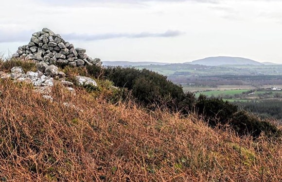 A small cairn on a hill with dry vegetation, distant hills, and a cloudy sky.