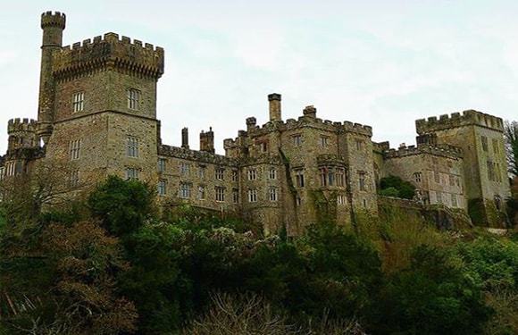 An image of a historic stone castle with towers and battlements, surrounded by greenery.