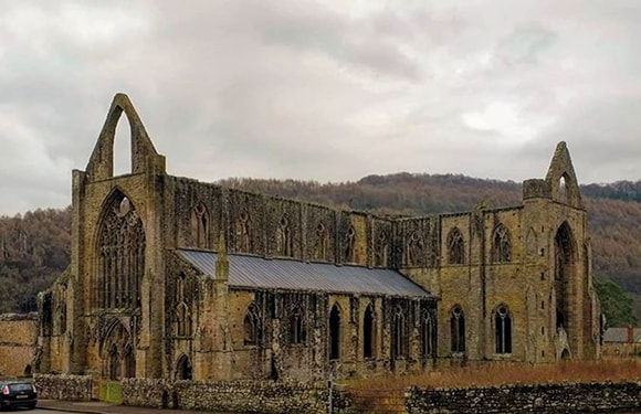 An image of the ruins of a large Gothic-style abbey with pointed arches and no roof, set against a backdrop of hills and a cloudy sky.