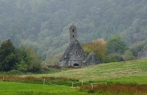 A small, ancient stone church with a distinctive bell tower, surrounded by lush greenery and set against a backdrop of misty, rolling hills.