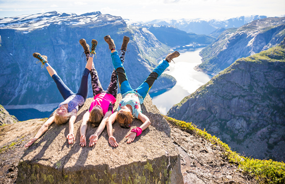 People lie on a rocky outcrop, legs raised, overlooking a river in a mountainous landscape.