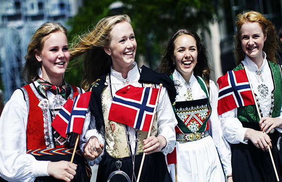 Four women smiling and walking, dressed in traditional Norwegian bunads with Norwegian flags, suggesting a celebration or national event.