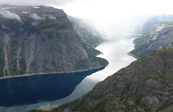 Aerial view of a fjord with steep cliffs on either side and a narrow body of water running through the center, under an overcast sky.