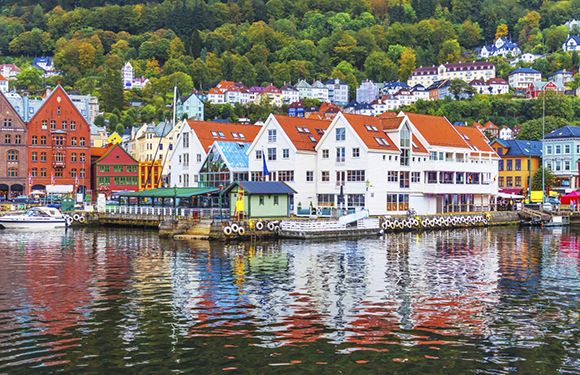 A colorful waterfront with a row of traditional buildings reflecting in the water, with a backdrop of green hills dotted with houses.