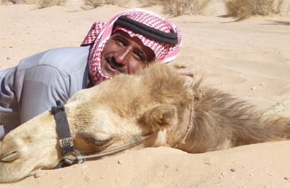 A man in traditional Middle Eastern attire lying next to a camel in a desert setting.