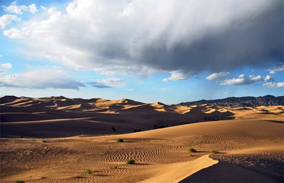 A landscape of patterned sand dunes under a partly clear, partly cloudy sky, hinting at changing weather.