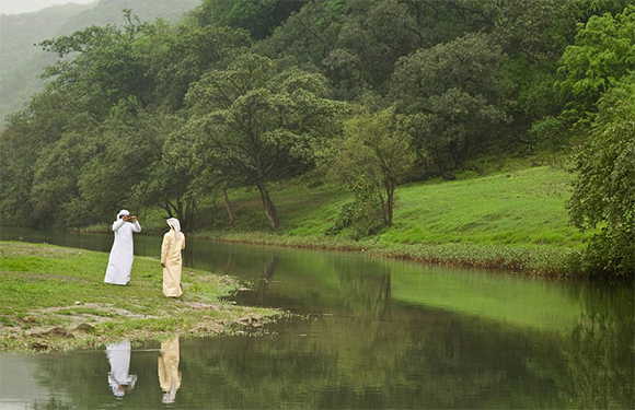 Two individuals in traditional clothes stand by a serene lake, their reflections and the surrounding greenery mirrored in the water.