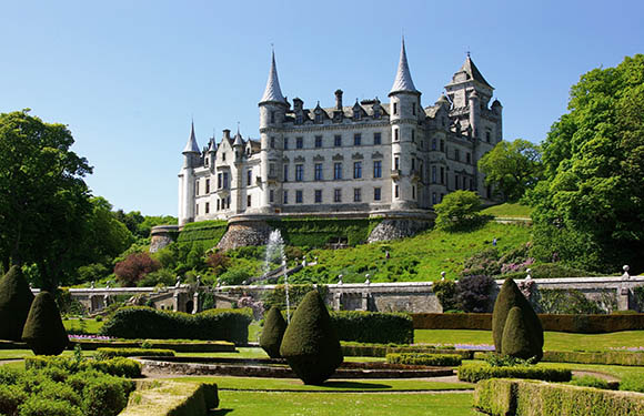 A picturesque castle with multiple spires set on a hill surrounded by manicured gardens, with a fountain in the foreground, under a clear blue sky.