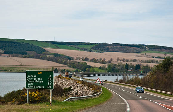 A car driving on a road with a directional sign on the left, overlooking a bridge crossing a body of water with hills in the background.