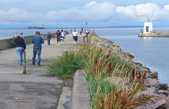 A group of people walking and jogging along a seaside promenade with a small lighthouse in the distance and overcast skies above.