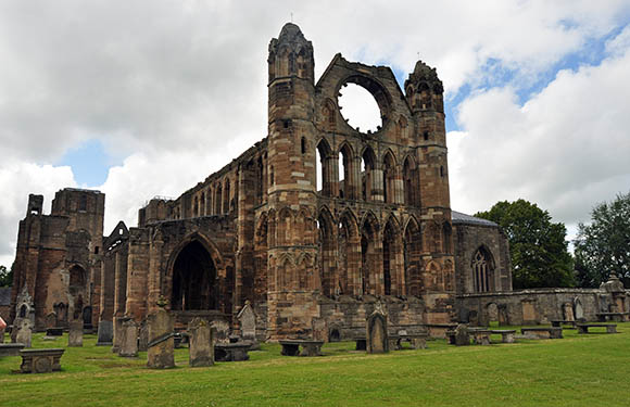 A photo of the ruins of an old Gothic church with an arched window, under a cloudy sky, surrounded by a grassy area with gravestones.