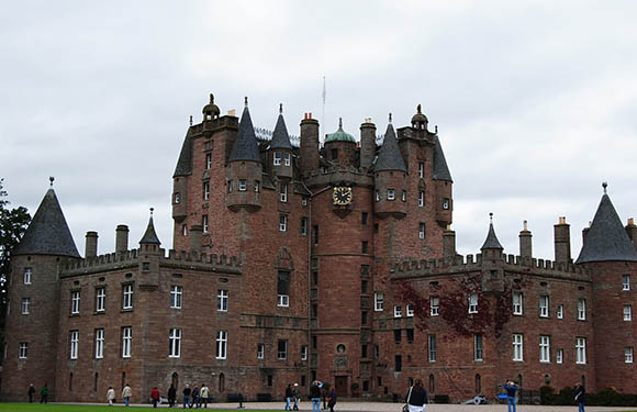 A historic castle with red and gray stonework, multiple spires, and turrets under an overcast sky, with people indicating its scale.