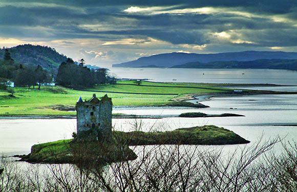 A small castle ruin on a grassy islet, surrounded by calm water, rolling hills, and a softly lit cloudy sky.