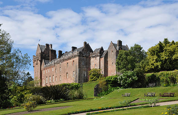 A historic castle with multiple spires set against a blue sky with clouds, surrounded by lush greenery and a manicured garden with benches.