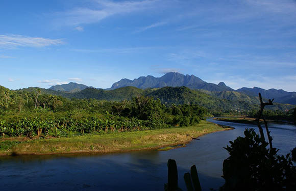 A picturesque scene with a river, lush greenery, mountains in the backdrop, and a blue, slightly cloudy sky.