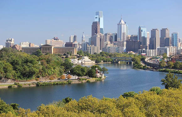 A daytime cityscape with a river, green banks, various buildings in the background, and a clear blue sky.