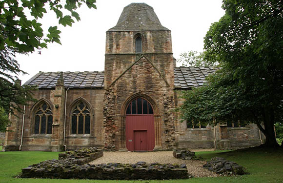 A small, traditional stone church with a red door, arched windows, and surrounded by green grass and trees.