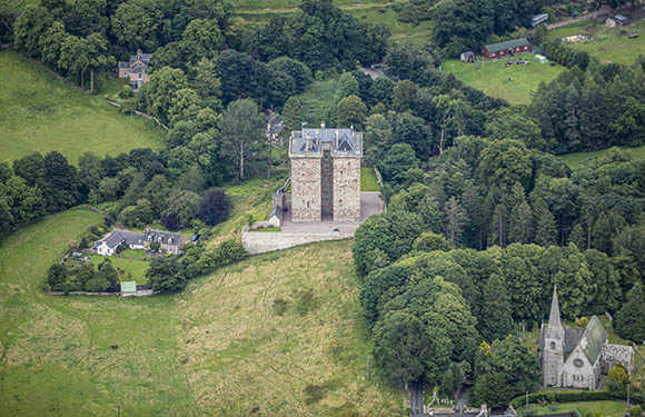 Aerial view of a historic stone tower surrounded by trees with a nearby church and green fields.