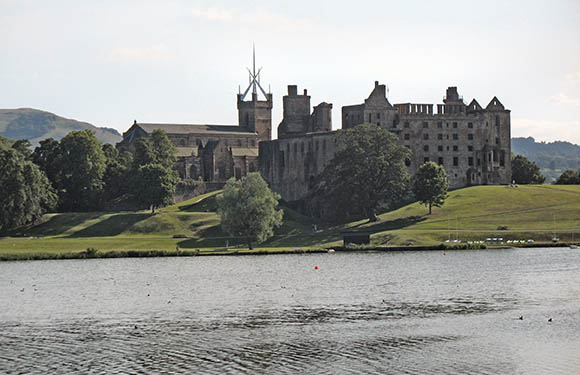 A castle with multiple towers and windows overlooking a body of water with trees and grass in the foreground under a clear sky with few clouds.