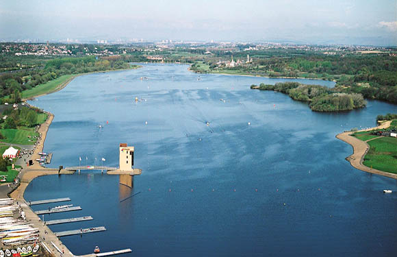 Aerial view of a large body of water with a pier and several boats, surrounded by greenery with a city skyline in the distance.